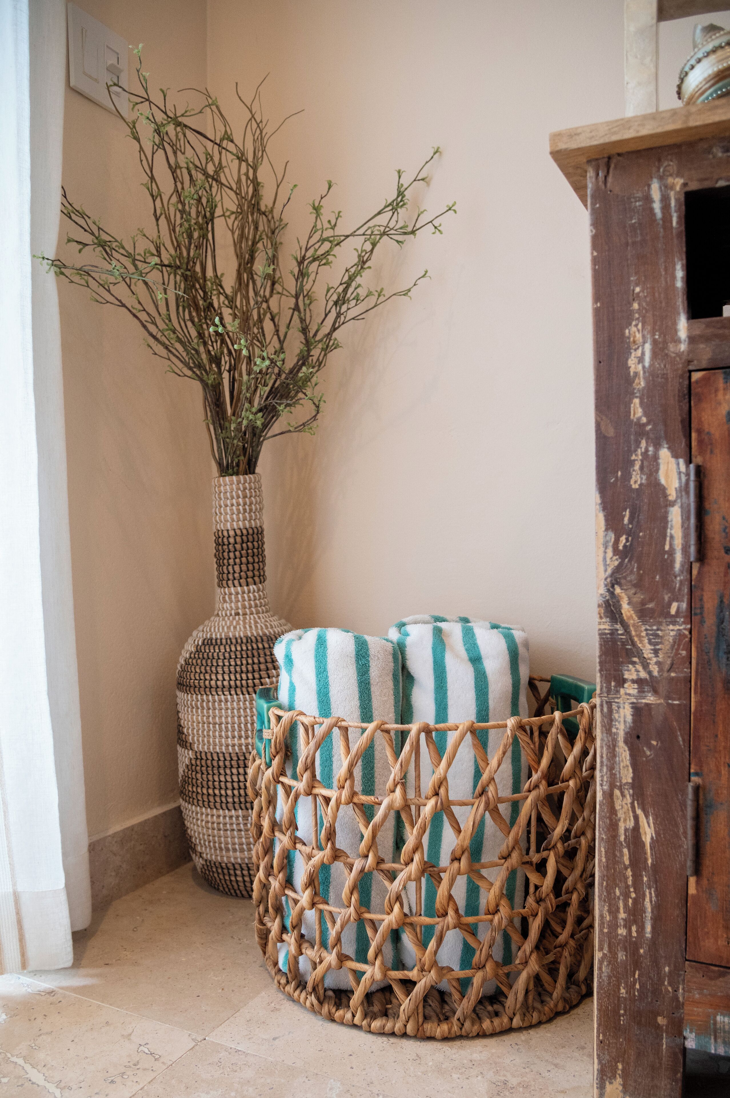 Un rincón con una cesta tejida, un jarrón alto con ramas y un mueble de madera.