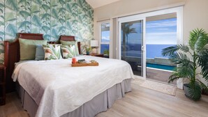 Une chambre avec un grand lit, une table de chevet, une commode, un plateau contenant divers objets, une vue sur l’océan par une porte coulissante et une plante en pot.