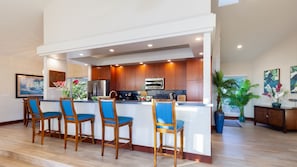A modern kitchen with wooden cabinets, stainless steel appliances, and a central island with bar stools. - NOV '26 Available! - INCREDIBLE 220 DEGREE OCEAN VIEW VILLA, SPA & FOUNTAIN POOL (Kamuela)