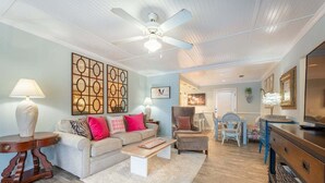 A living room with a ceiling fan, a sofa, a coffee table, a TV, and a dining area with a chair and table. - Direct Ocean View 2BR Condo Near Pier Village | St. Simons Island Rental (St. Simons Island)