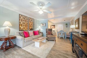 A living room with a ceiling fan, a sofa, a coffee table, a TV, and a dining area with a chair and table. - Direct Ocean View 2BR Condo Near Pier Village | St. Simons Island Rental (St. Simons Island)