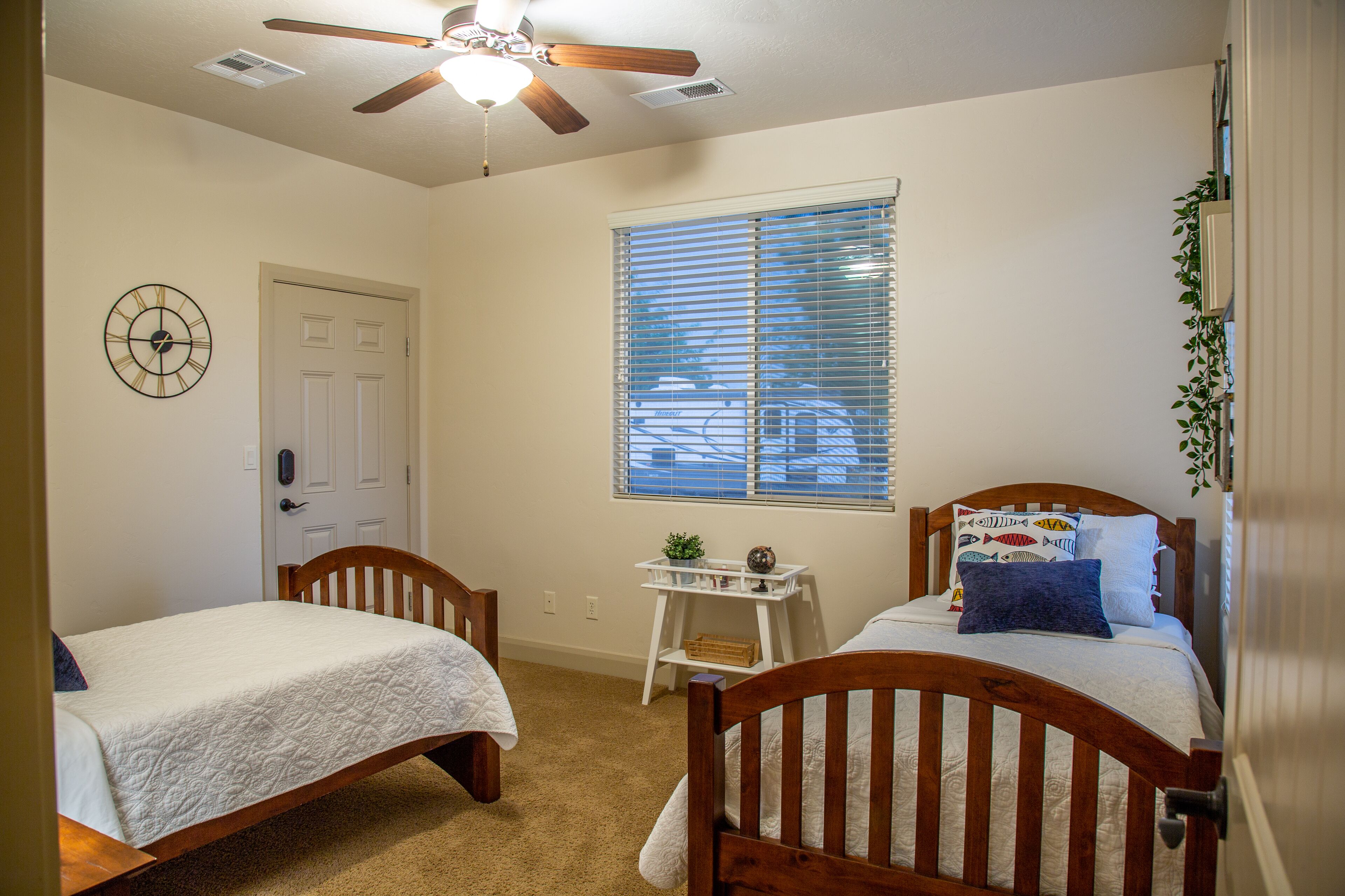 A bedroom with two wooden beds, a ceiling fan, a window with blinds, a clock, and a small table with a plant.