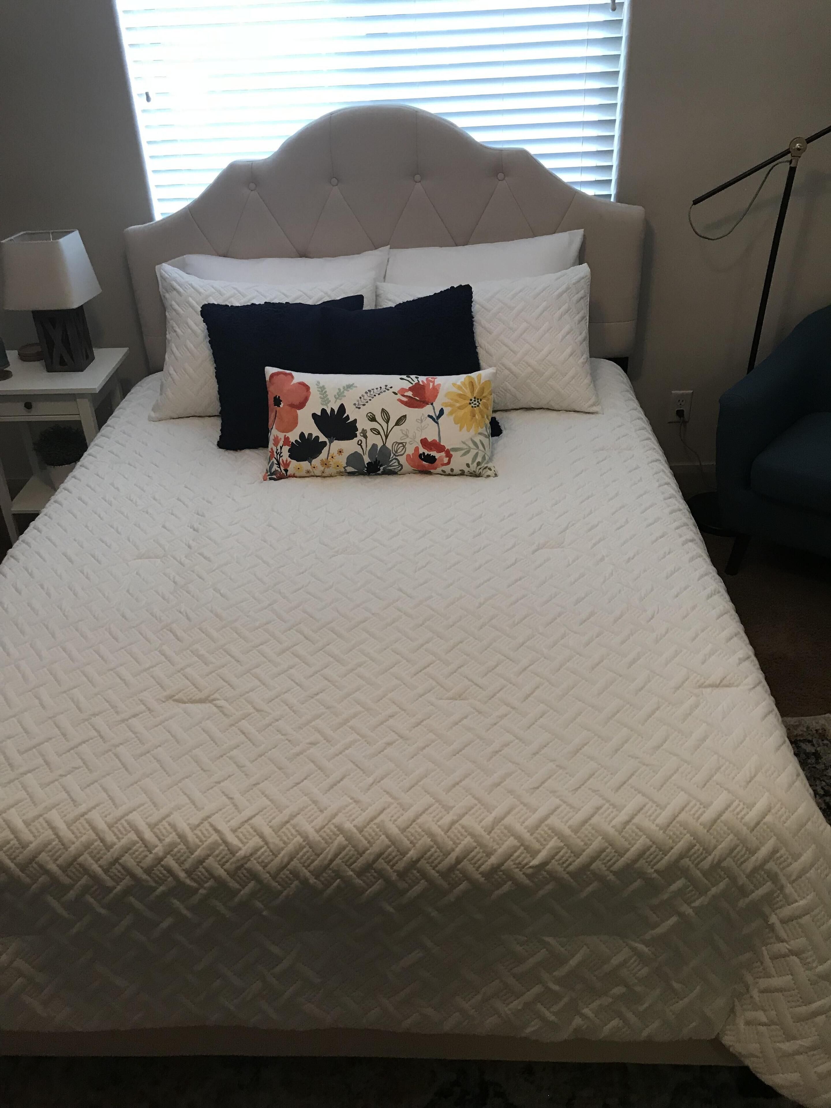 Un lit bien fait, avec une couette blanche matelassée, un coussin à motifs floraux et un coussin bleu foncé.