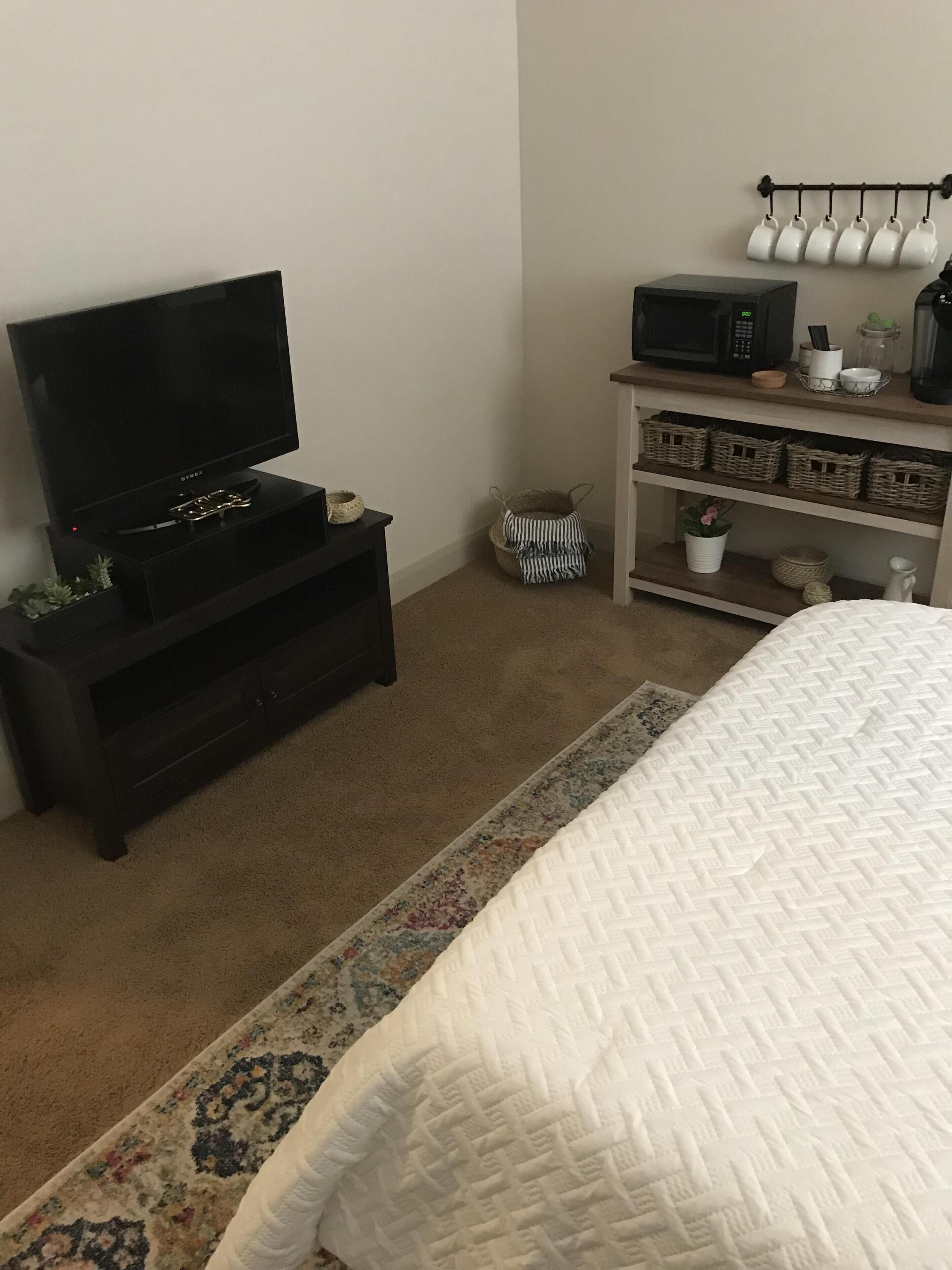 Une chambre à coucher avec un lit, un téléviseur posé sur un meuble, un four à micro-ondes et une étagère avec des paniers et une plante.