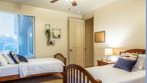 A bedroom with two single beds, a ceiling fan, a window with blinds, a door, a bedside table with a lamp, and a wall-mounted mirror.