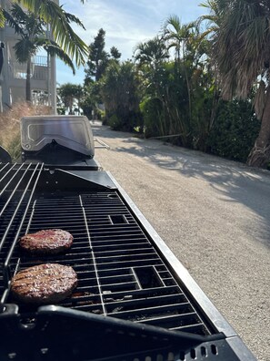 Ein Grill mit zwei Bratburgern, umgeben von Palmen und einem Gebäude im Hintergrund.