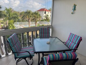 Un balcon avec une table en verre et des chaises Ă rayures, donnant sur une piscine et des immeubles.