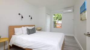 A neatly arranged bedroom with a wooden bed, white bedding, two bedside lamps, a small table, and a window with a view of greenery. - Close to beach, downtown, airport. Entire Apt. (Fort Lauderdale)