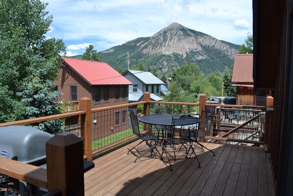 A wooden deck with a table and chairs, overlooking a mountain and a red-roofed building.