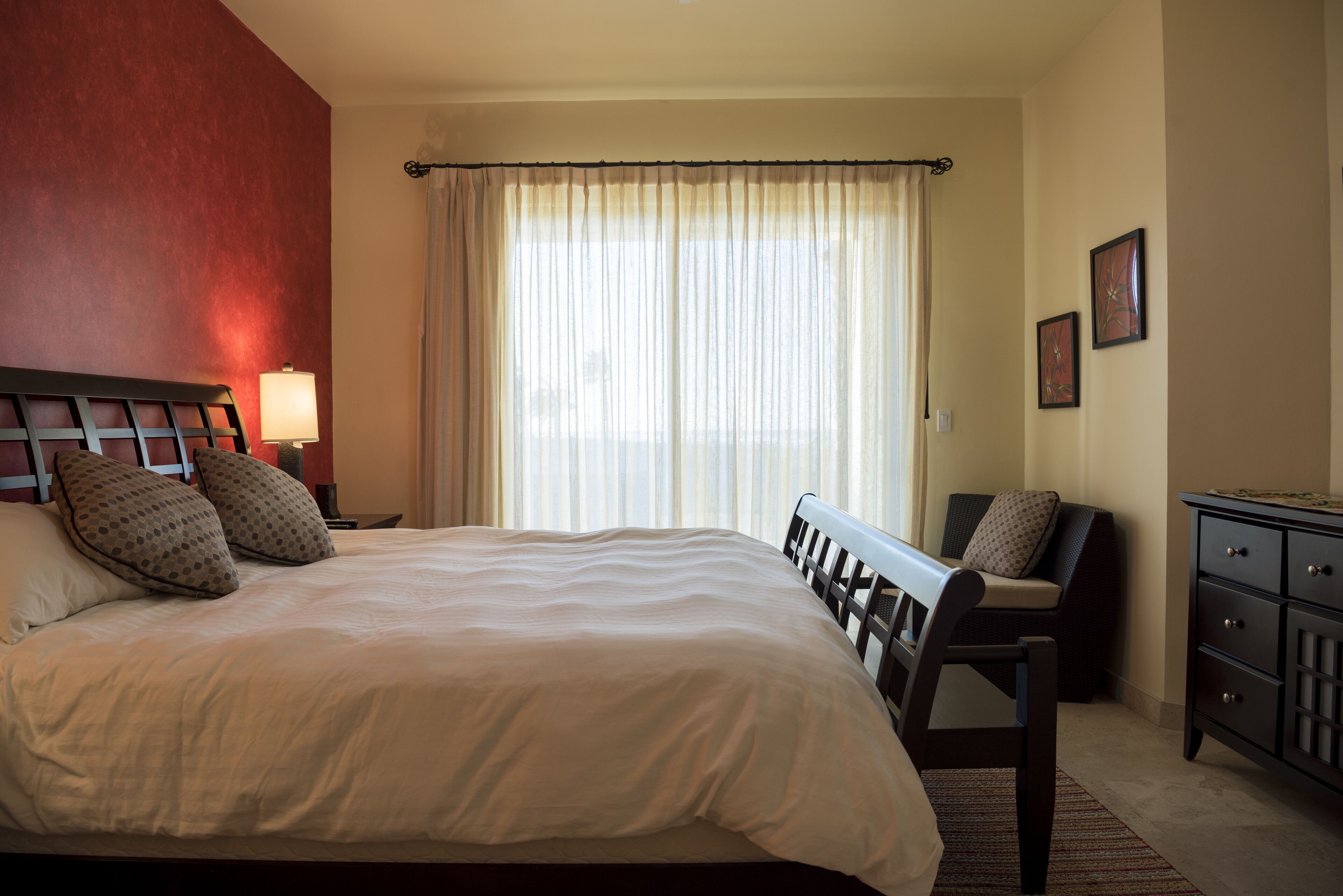 Quarto de hotel com uma cama grande, mesa de cabeceira com abajur, uma cadeira, cômoda e janela com cortinas.