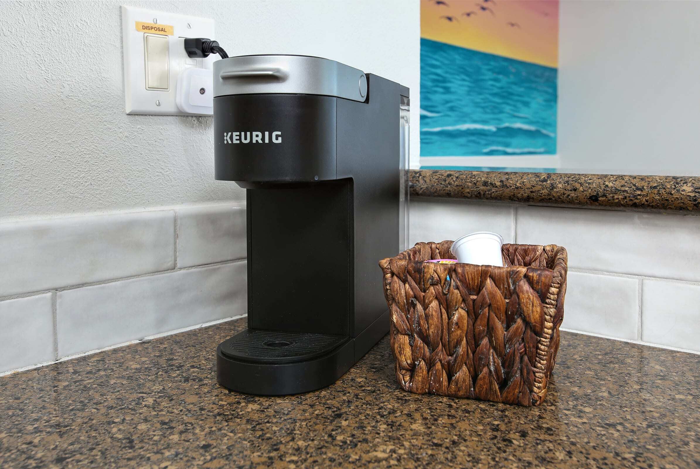 Una cafetera Keurig sobre una encimera de granito con un cubo de basura al lado.