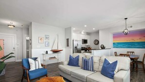 A modern living room with a sofa, armchairs, and a coffee table. There is a kitchen area with a refrigerator and microwave. A painting of a sunset over the ocean is on the wall.
