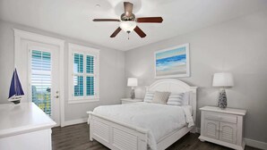 A bedroom with a white bed, two nightstands, a ceiling fan, and a window with shutters.