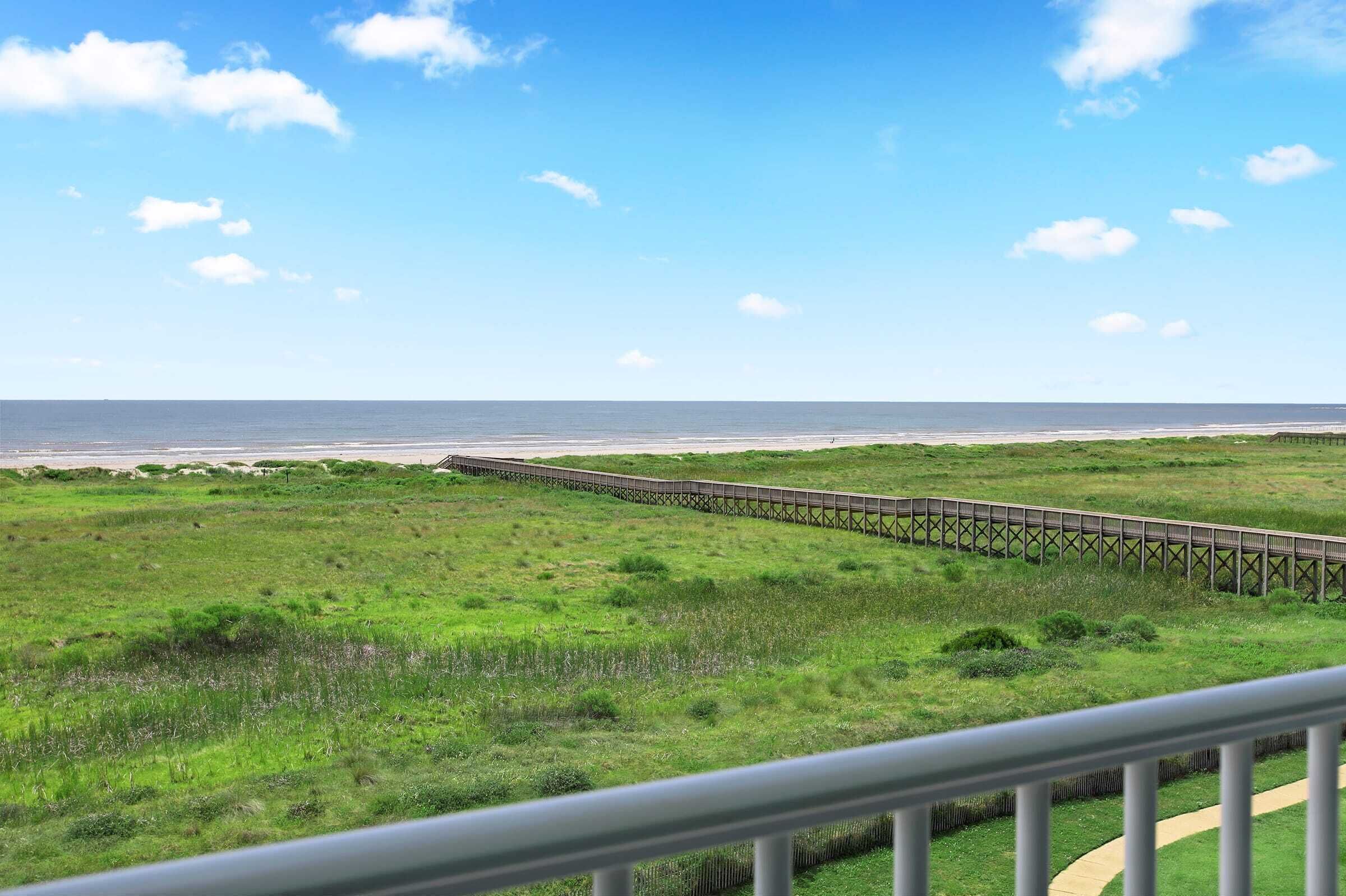 A coastal landscape with a wooden boardwalk extending towards the ocean, surrounded by grassy fields.