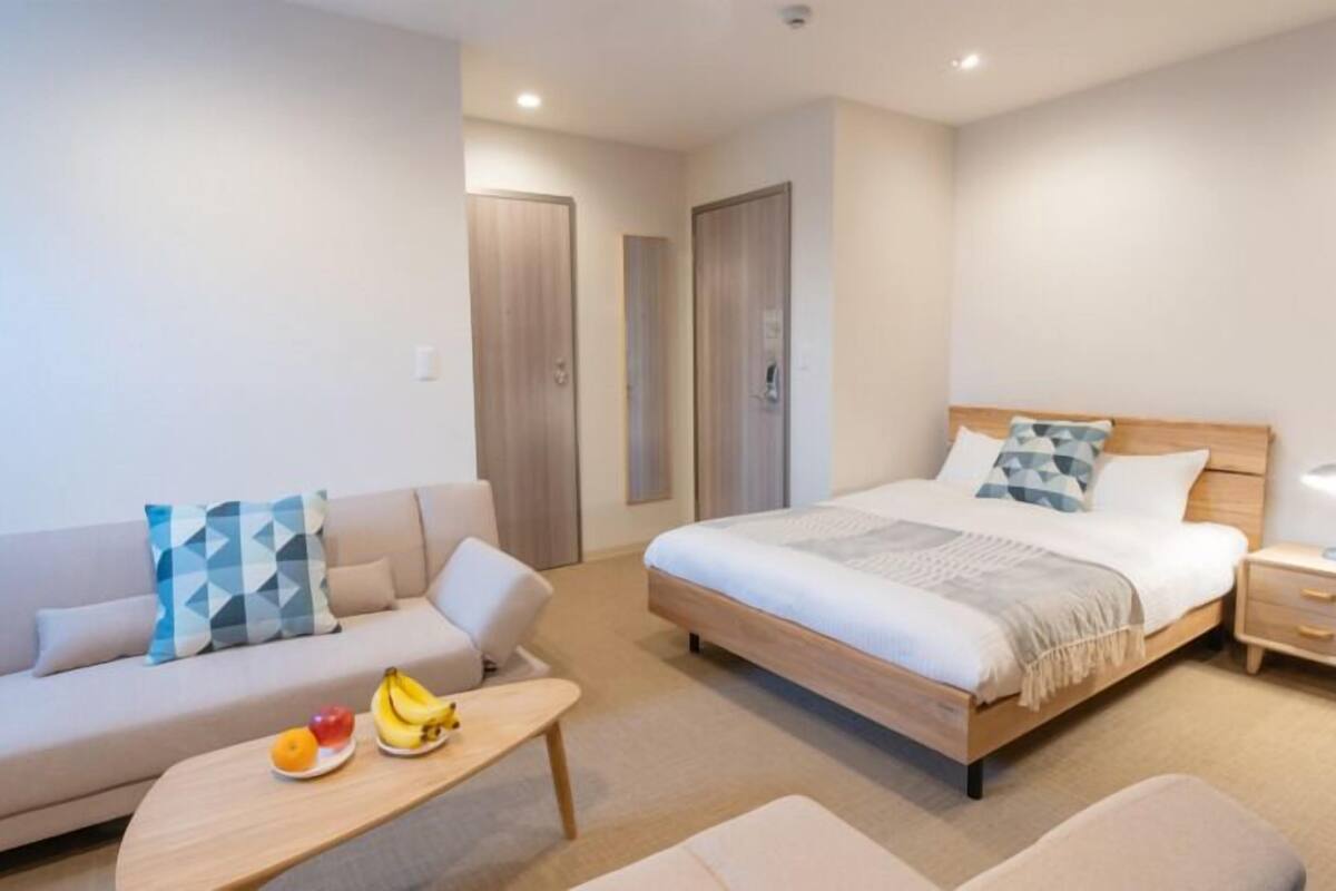 A hotel room with a bed, a sofa, a small table with fruit, a nightstand with a lamp, and a wardrobe.