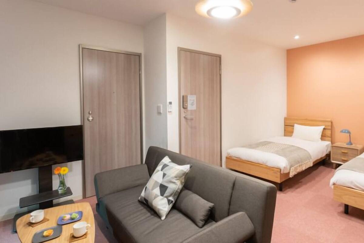 A hotel room with a sofa, two single beds, a TV, and a small table with cups and a vase.