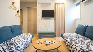 Ein modernes Hotelzimmer mit zwei blau gemusterten Sofas, einem Holztisch mit einer Speisenplatte, einem flachbildfernseher, der über einer Kommode angebracht ist, und einer Klimaanlage an der Wand.