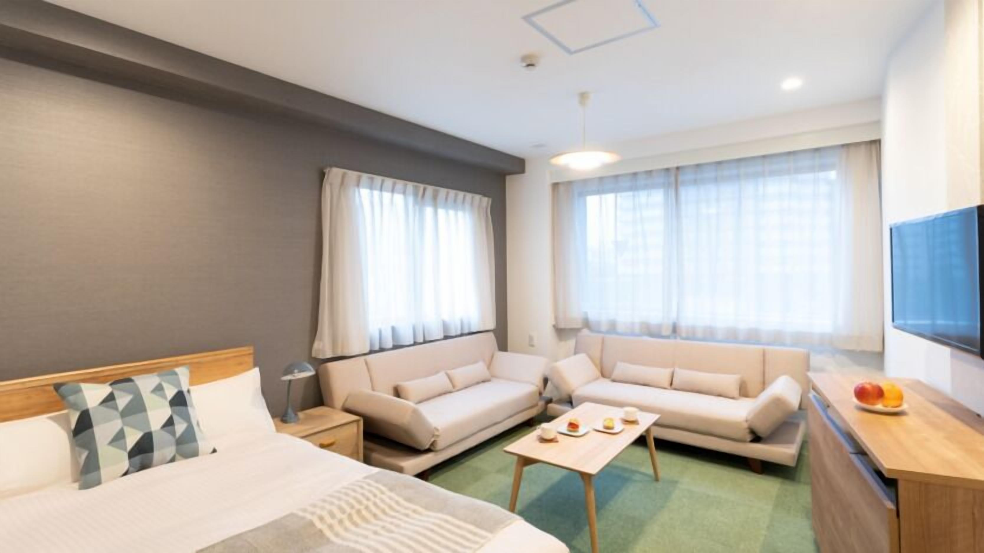 Habitació d'hotel amb un llit, dos sofàs, una taula petita, un televisor i una finestra amb cortines.