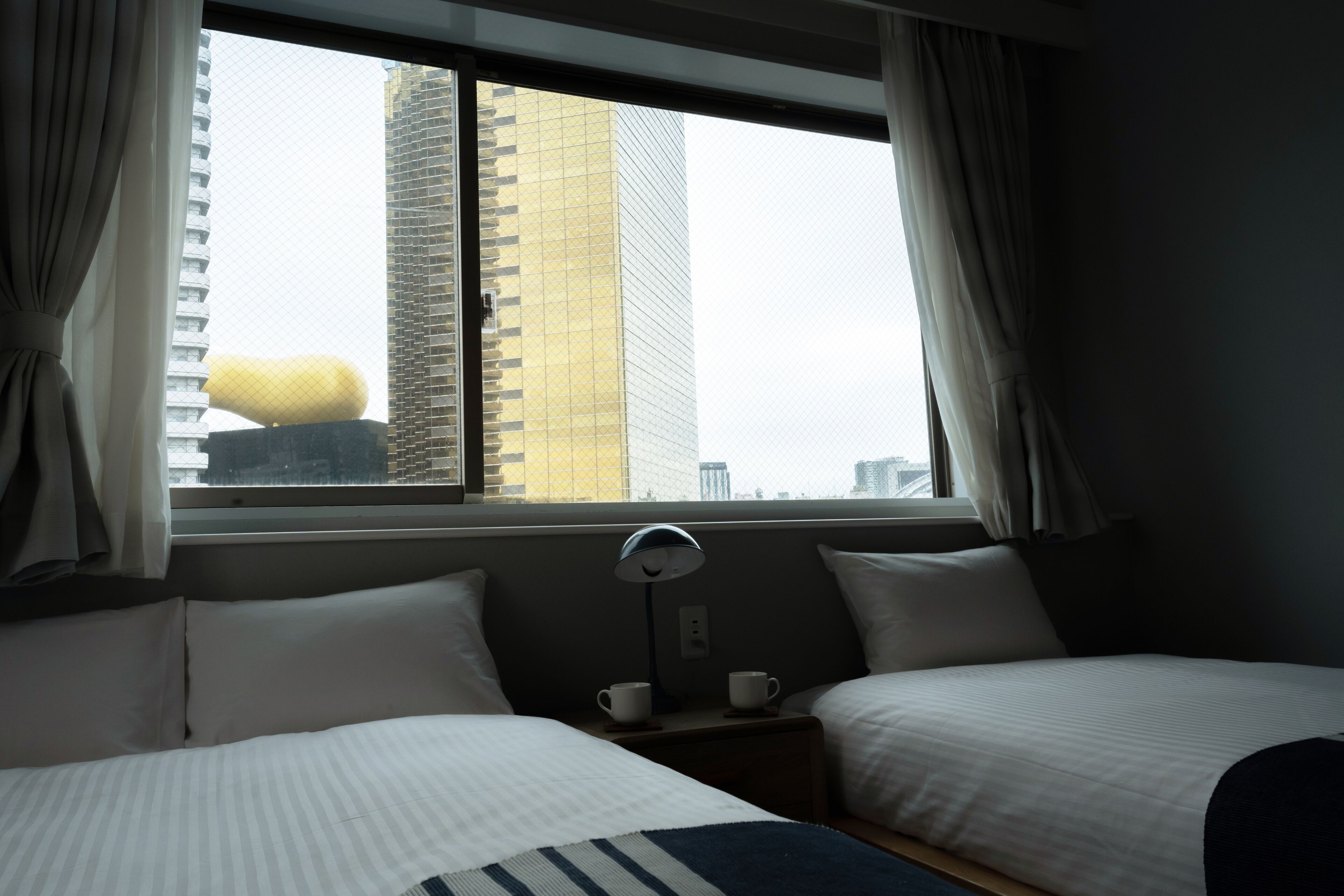A hotel room with two beds, a window with curtains, a bedside table with a lamp, and a view of tall buildings.