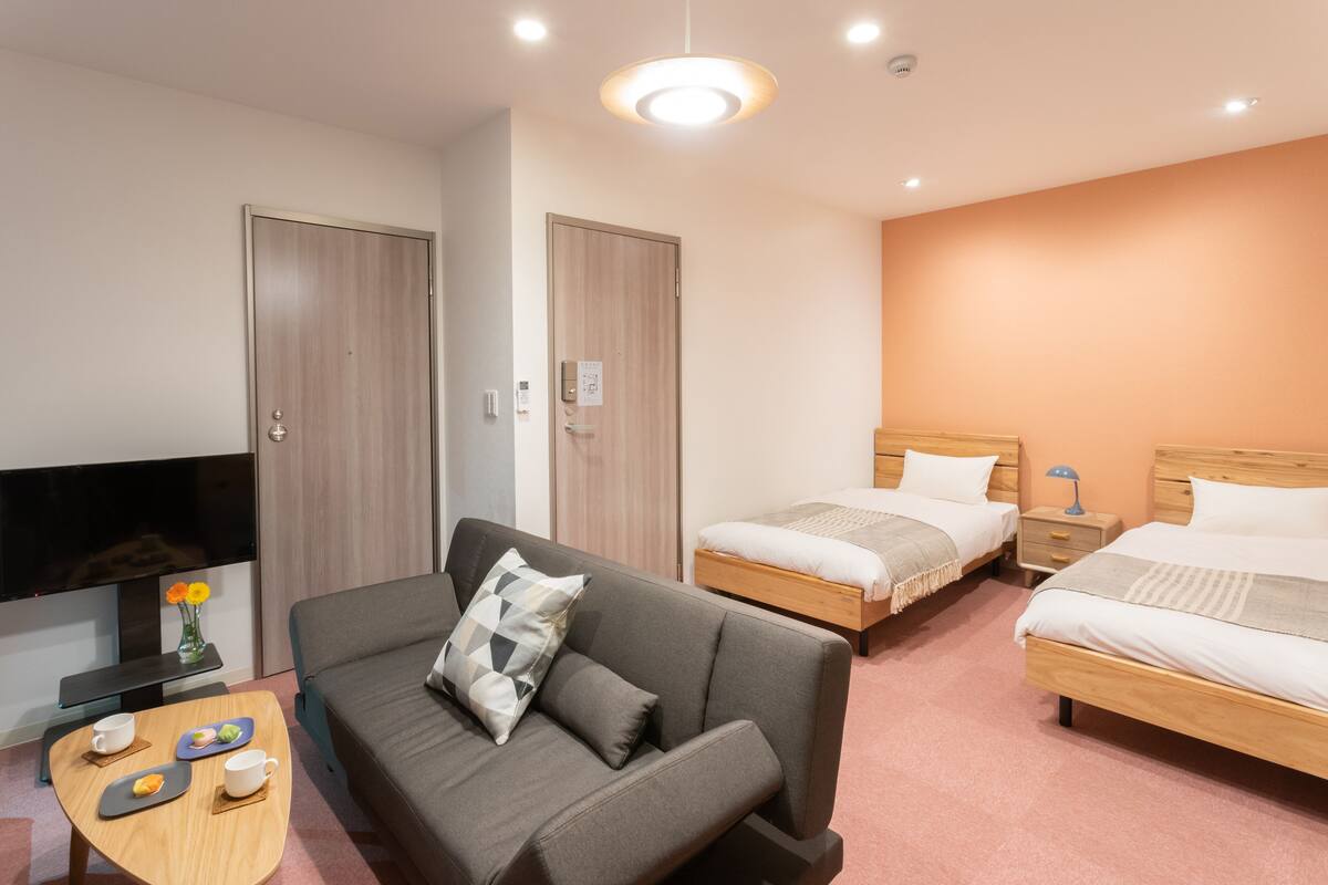 A hotel room with a sofa, two single beds, a TV, and a small table with cups and a vase of flowers.