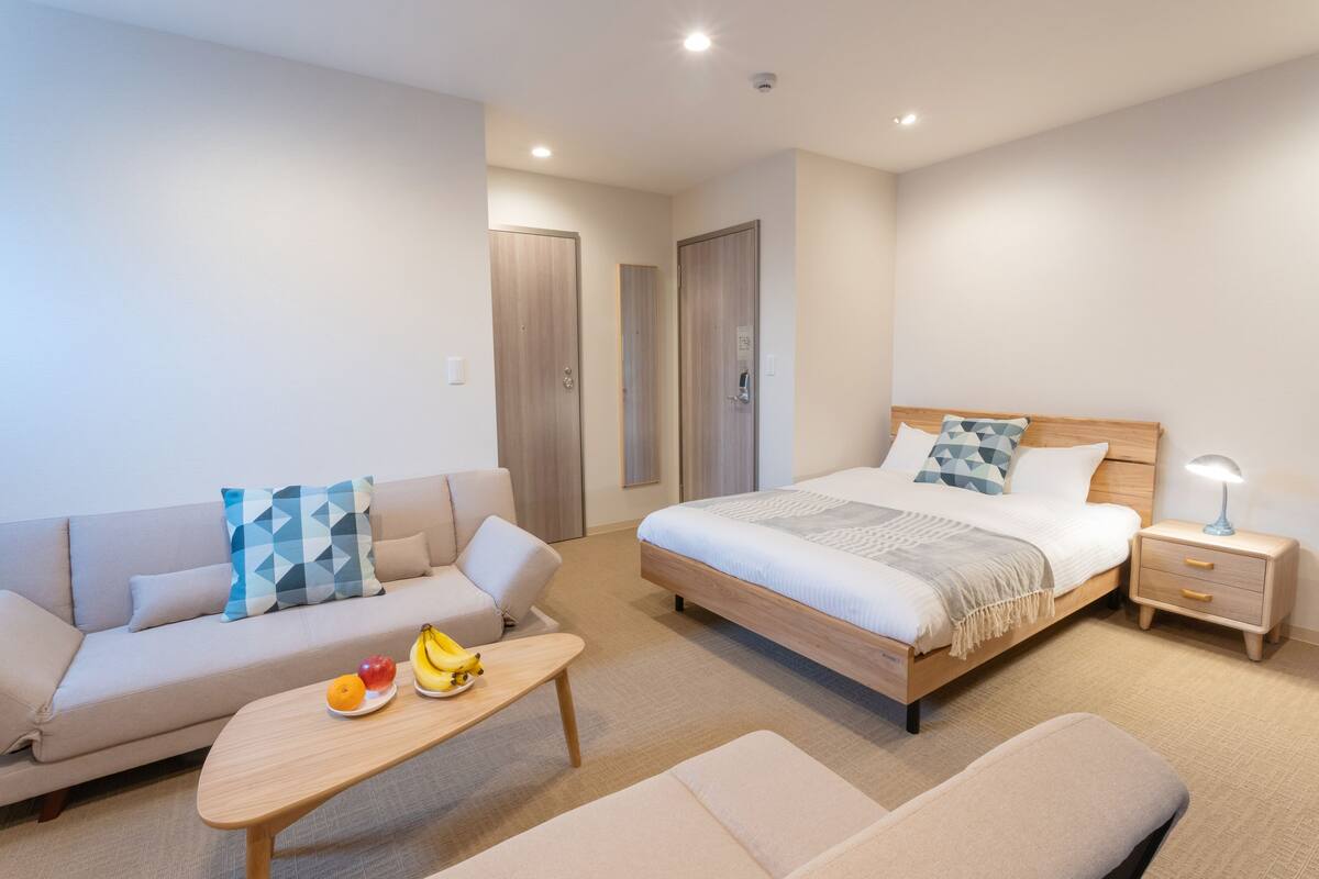 A modern hotel room with a bed, a sofa, a small table with fruits, and a bedside lamp.