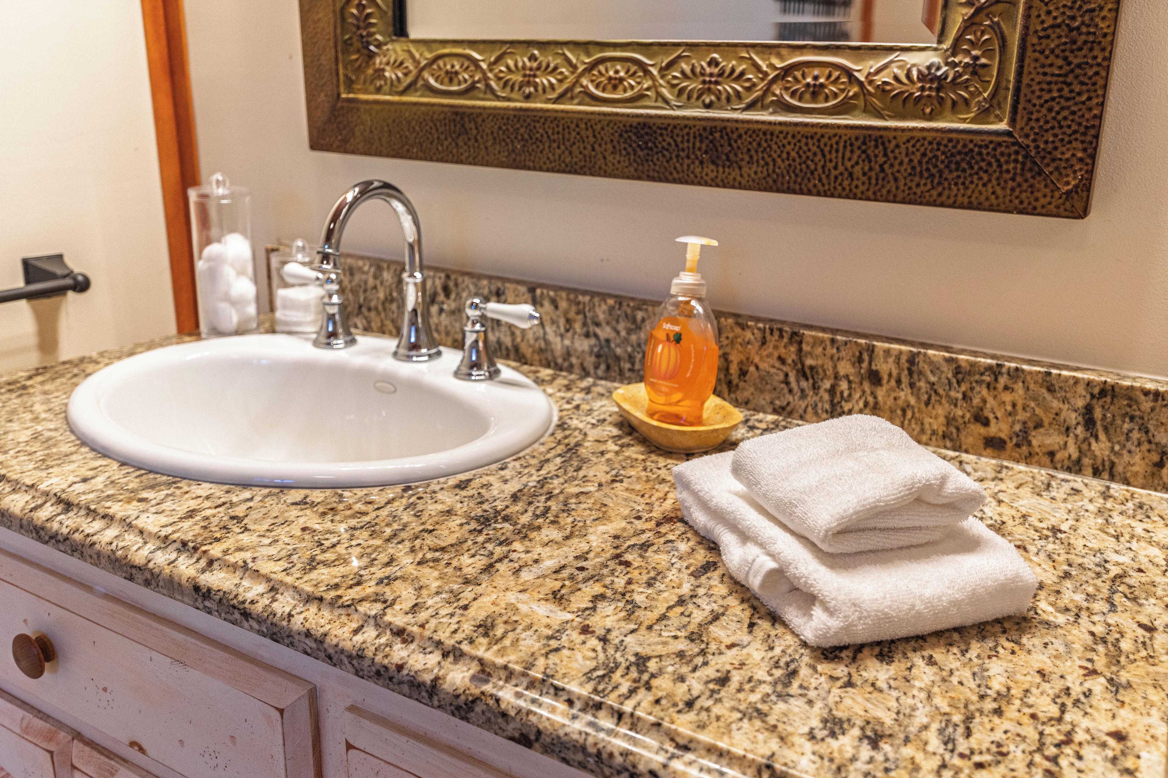 A bathroom with a granite countertop, a white sink, a mirror with an ornate frame, a soap dispenser, and a stack of white towels.