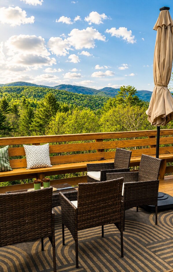An outdoor seating area with wicker chairs and a wooden bench, overlooking a forested landscape with hills.
