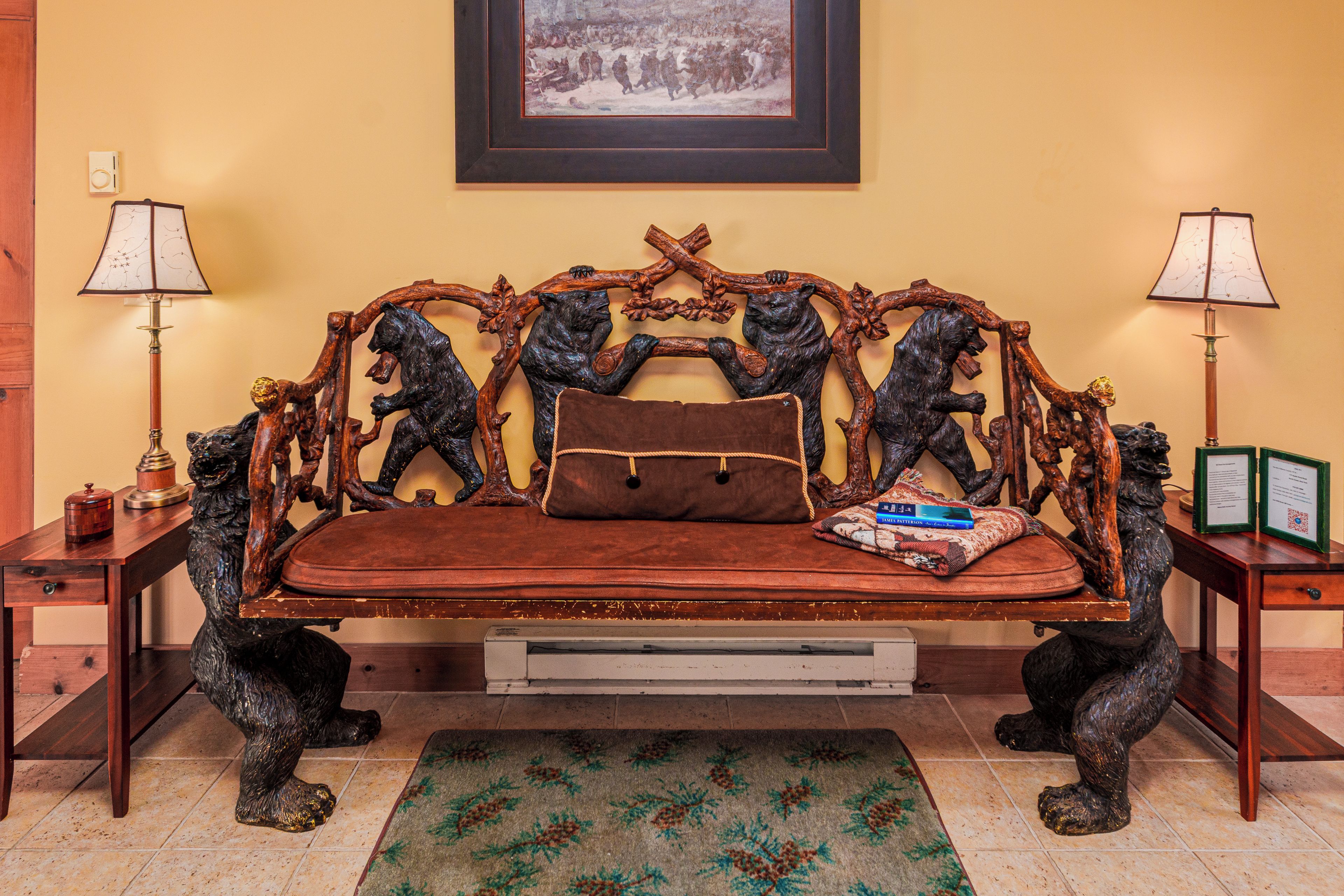 Un canapé doté d’un coussin en cuir marron, flanqué de deux accoudoirs en bois ornés de sculptures d’animaux, un tableau encadré au mur et deux lampes de chaque côté.