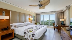 Una camera d'albergo con un letto grande, una scrivania con una sedia, una televisione, una ventola a soffitto e un balcone con vista.