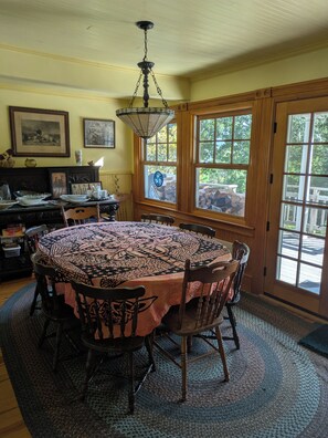 A dining room with a round table, chairs, a chandelier, framed artwork, and a view through large windows.