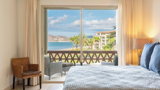 A bedroom with a large bed, a chair, and a view of the beach and palm trees. - E303-Beautiful front ocean condo (La Paz)