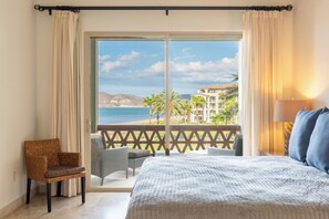 A bedroom with a large bed, a chair, and a view of the beach and palm trees.