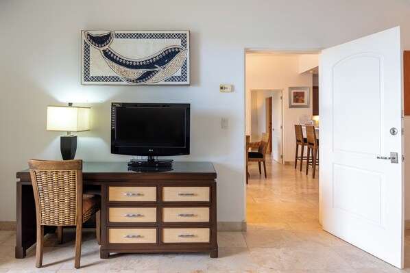 A hotel room with a TV on a wooden cabinet, a wicker chair, and a dining area in the background.