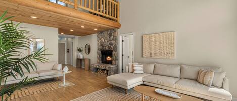 A spacious living room with a high ceiling, a stone fireplace, a wooden balcony, and a large window.