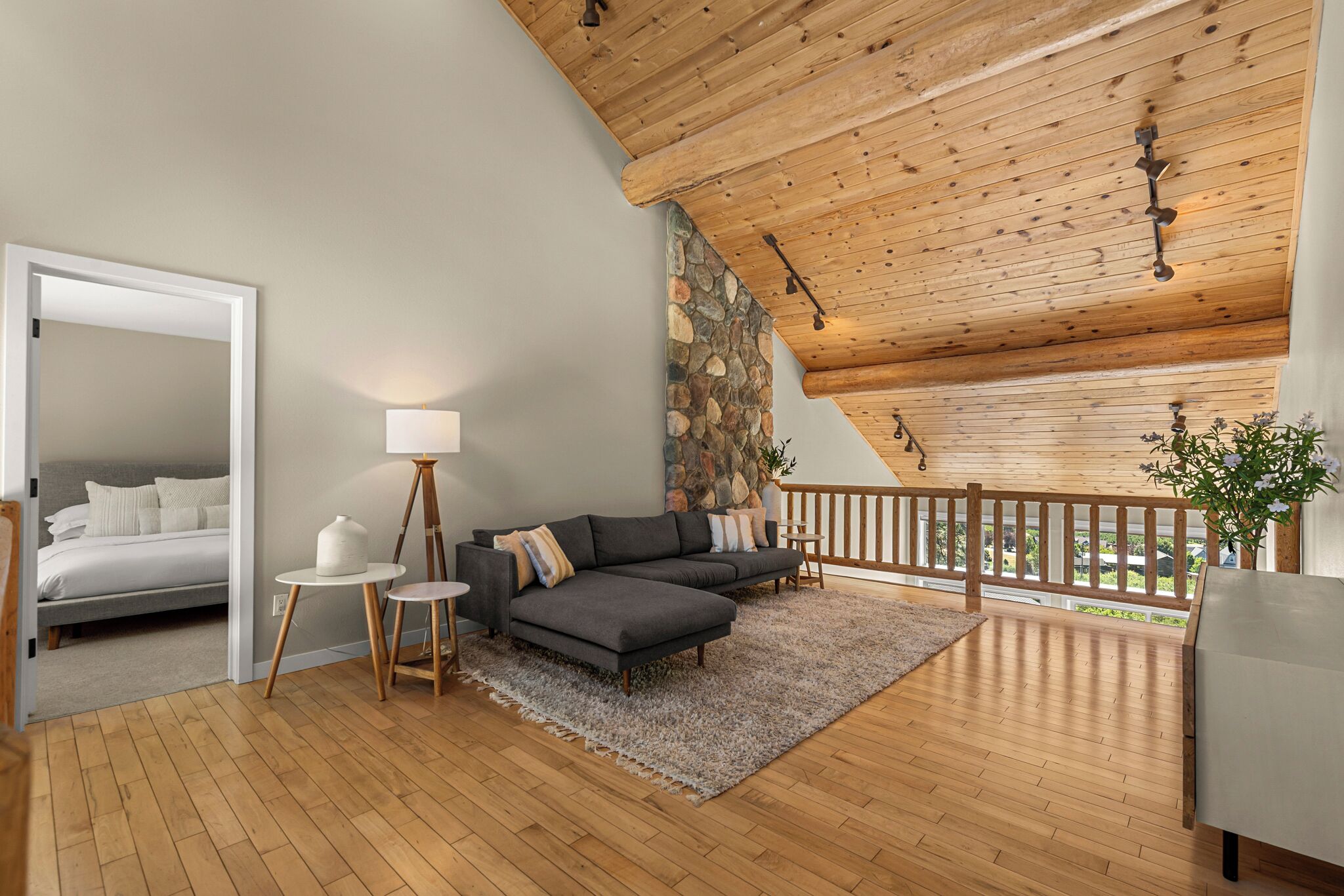 Een modern interieur met een houten plafond, een stenen muur, een grijze bank, een klein rond tafeltje en een bed in de aangrenzende kamer.