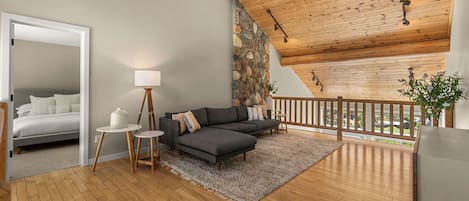 Een modern interieur met een houten plafond, een stenen muur, een grijze bank, een klein rond tafeltje en een bed in de aangrenzende kamer.