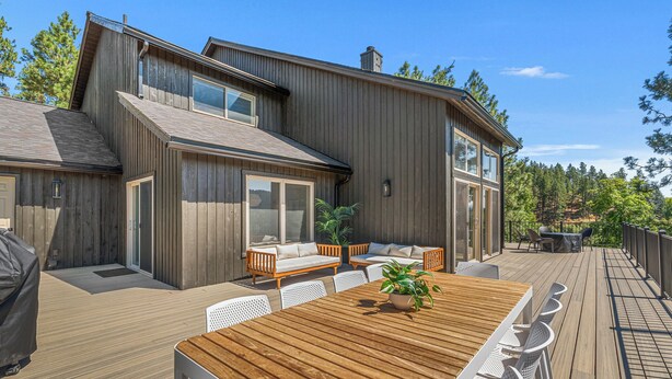 Eine moderne Holzterrasse mit Esstisch und Stühlen, einer integrierten Bank mit Kissen und Blick auf den umliegenden Wald.