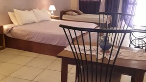 A bedroom with two beds, a dining table with chairs, a lamp, and a window with curtains. - Grazia Maria Hotel (Mbour)