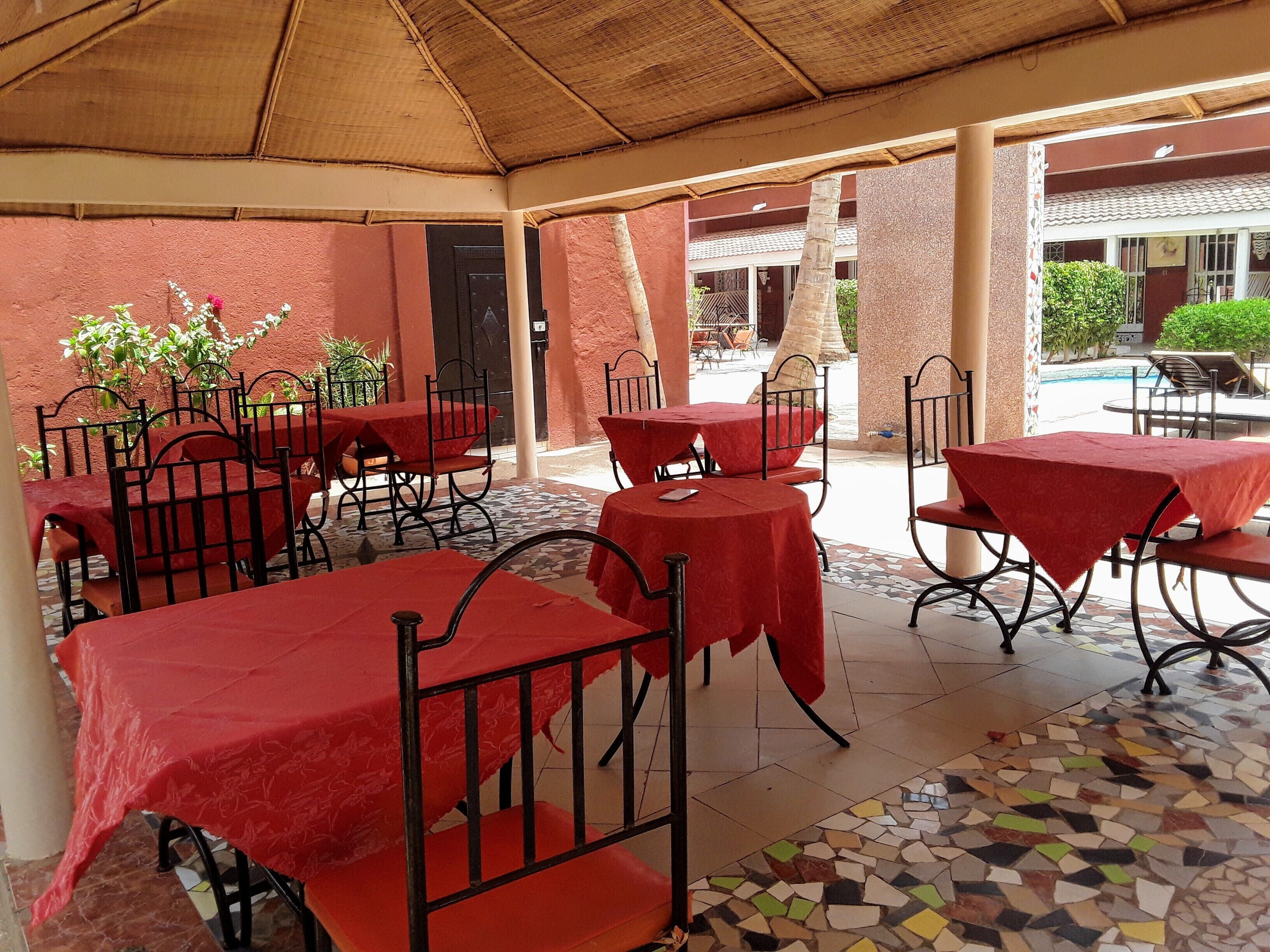 Un patio al aire libre con mesas y sillas cubiertas con manteles rojos, suelo de baldosas y techo de paja.