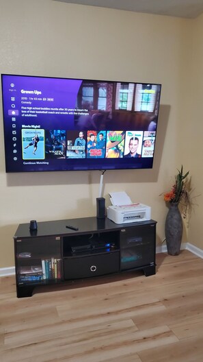 Un televisor de pantalla plana montado en la pared, sobre un mueble negro que alberga una impresora y un jarrón con flores.