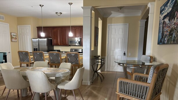 A dining area with a round table, chairs, and a glass-top table. There is a kitchen area with cabinets and appliances in the background.