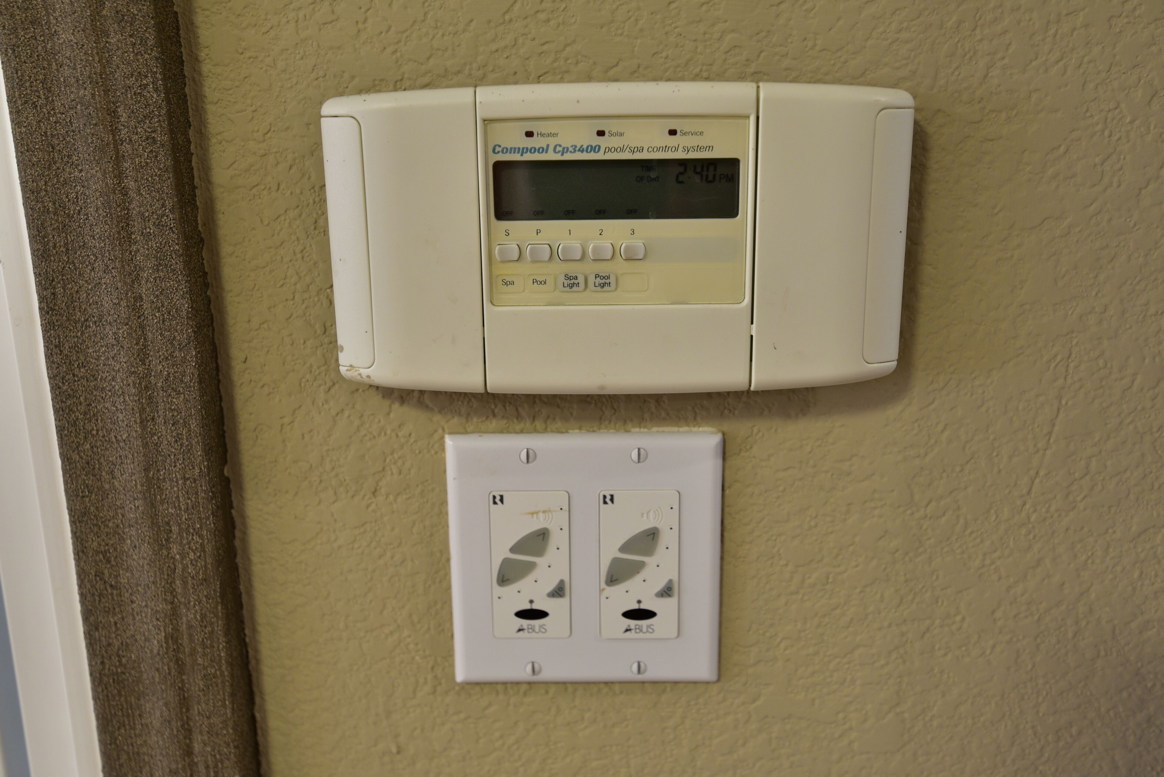 A control panel with a digital display for a pool/spa system, two switches below it, and a wall-mounted thermostat.