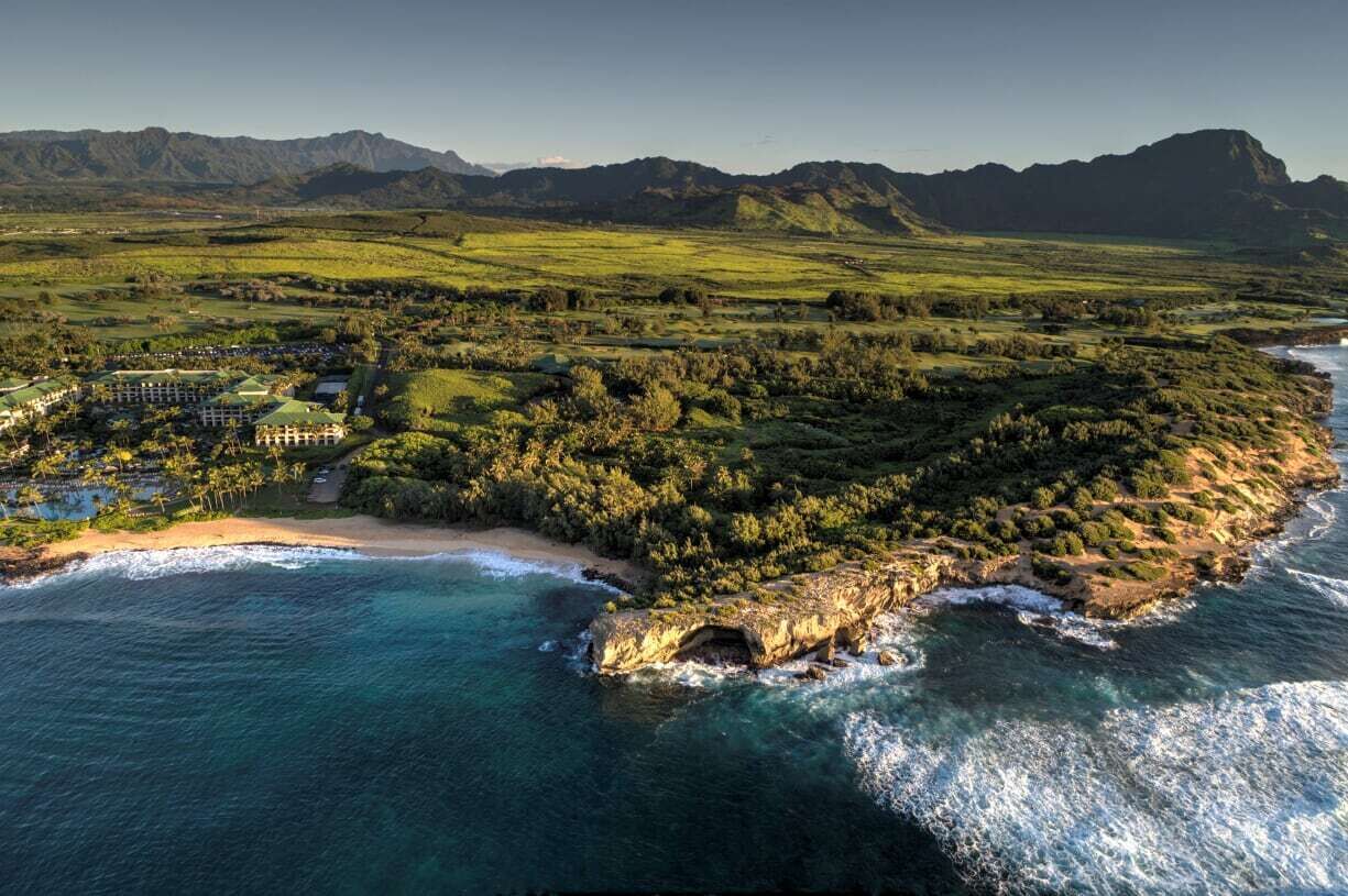 Vista aérea de un complejo turístico costero con playa, exuberante vegetación y montañas al fondo.