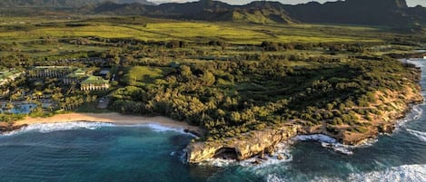 An aerial view of a coastal resort with a beachfront, lush greenery, and mountainous backdrop.