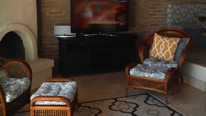 Un salon doté d’une cheminée en briques, d’un téléviseur à écran plat fixé au-dessus et de deux fauteuils en osier ornés de coussins à motifs.