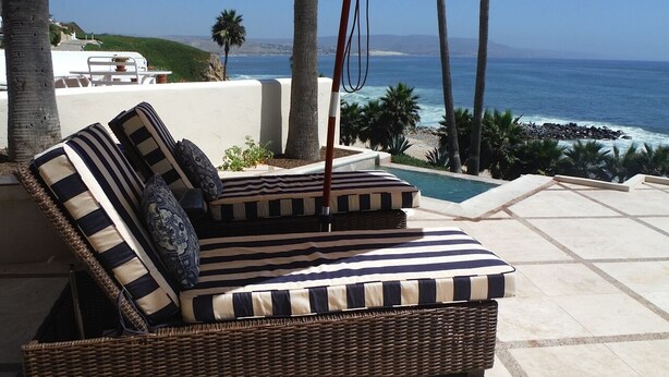 Un fauteuil de jardin en osier, doté de coussins à rayures et surplombé d’un parasol jaune, offre une vue sur la piscine et l’océan.