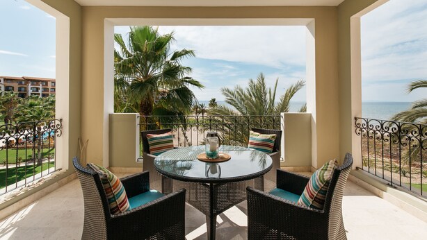 A balcony with wicker furniture, a glass table, and a view of the sea and palm trees. - Beguiling pool-ocean view unit (La Paz)