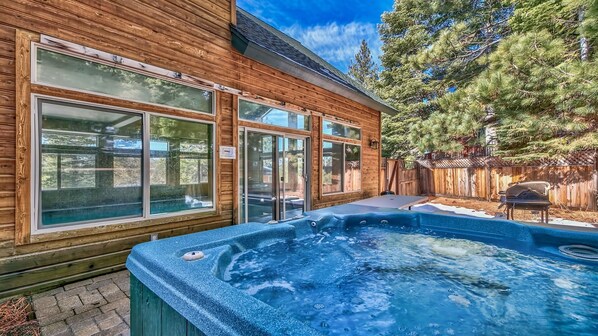 A wooden cabin with large windows, a hot tub, and a fenced backyard with trees.