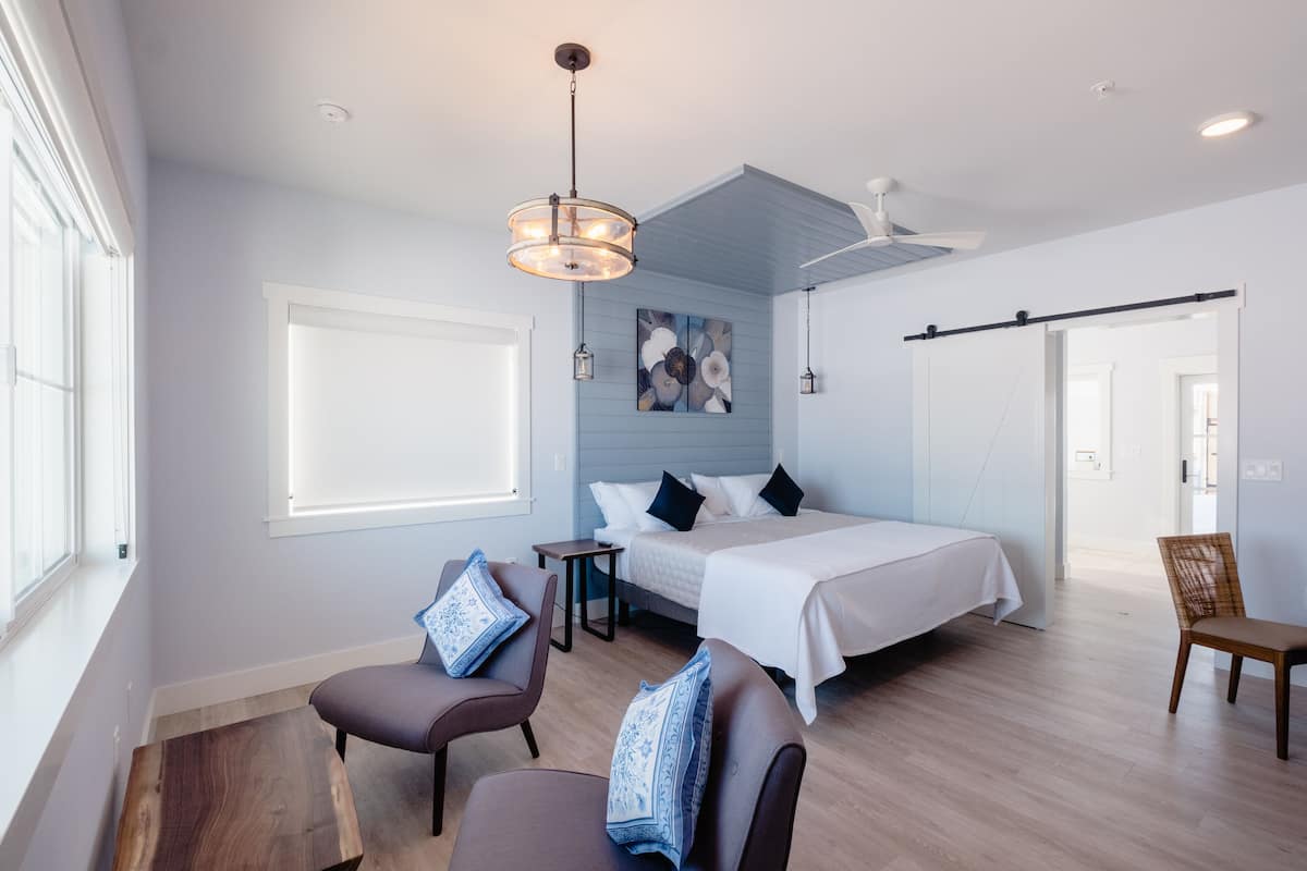 Un dormitorio moderno con una cama grande, un escritorio y una silla. Hay una ventana, un ventilador de techo y un cuadro en la pared.
