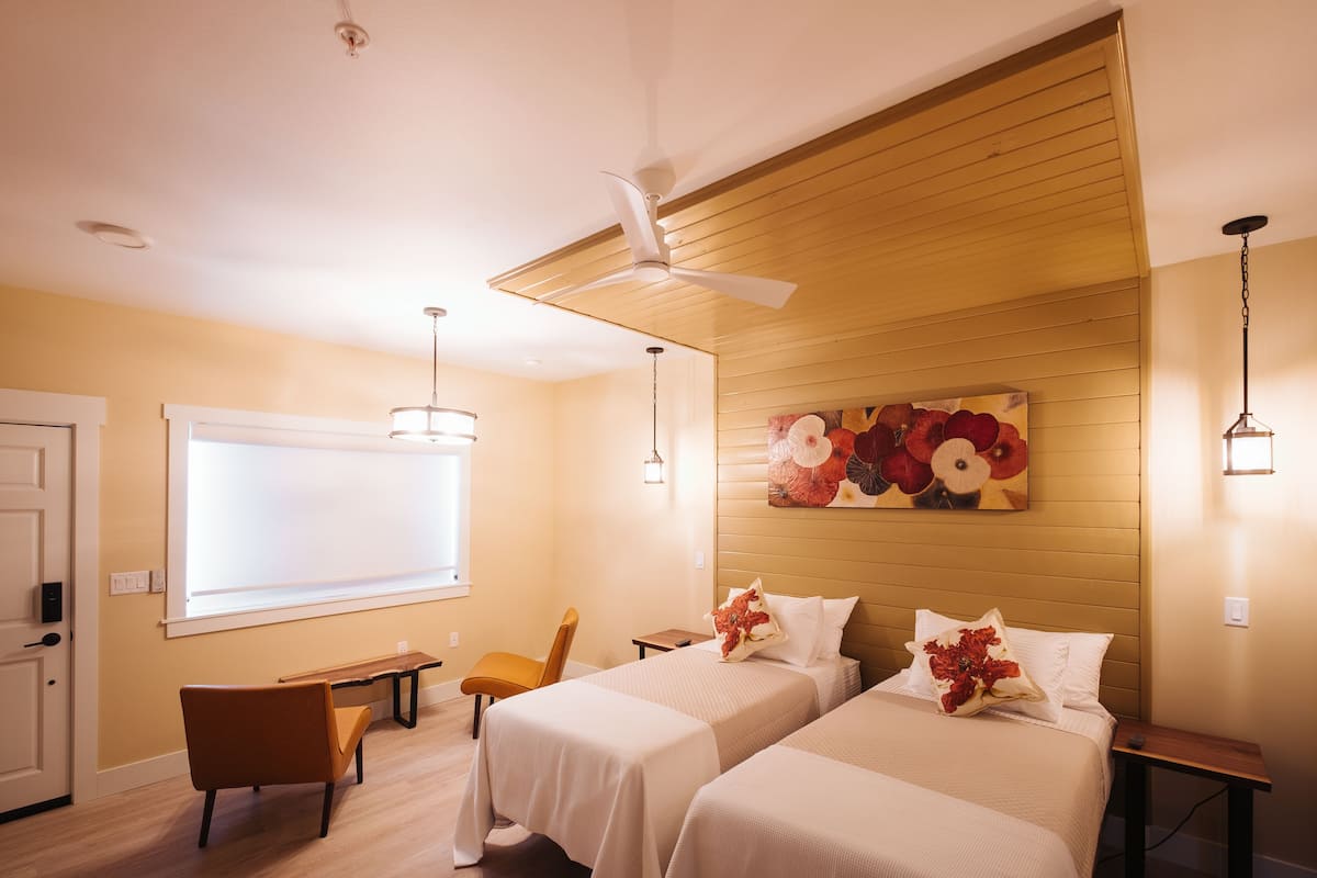 Habitación de hotel con dos camas, ventilador de techo, escritorio, una silla y un cuadro grande con motivos florales en la pared.